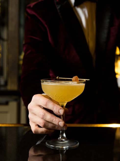 A bartender dressed in a velvet tuxedo serves an orange cocktail at a bar