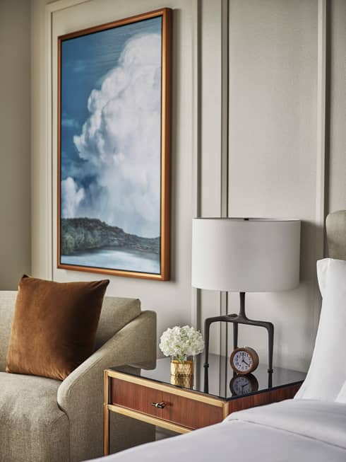 Framed painting of clouds over lake above armchair, nightstand with lamp, clock