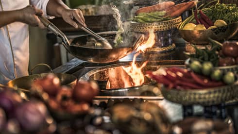 Close-up of chef stirring meals in copper pans over stove, flames 