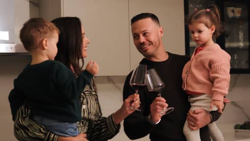 Two adults, each holding a small child in their arms, smile at each other and click their glasses of wine together while standing behind a kitchen counter