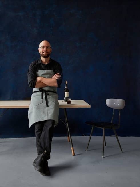 Portrait photo of Chef Greg Vernick leaning against table with bottle of wine