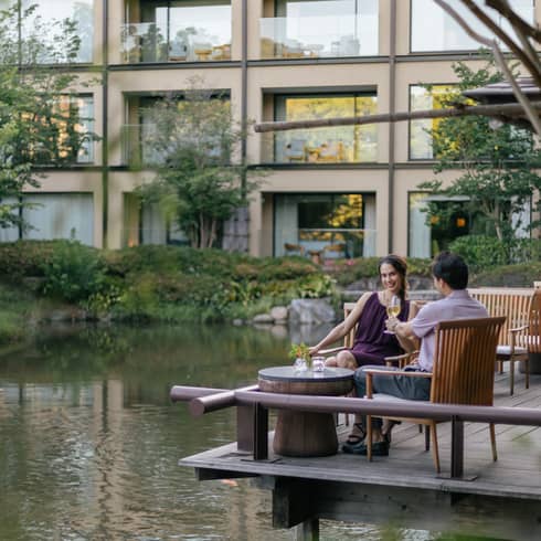 Smiling couple clink champagne flutes at a café table on a wood deck overlooking a rippling pond bordered by lush foliage.