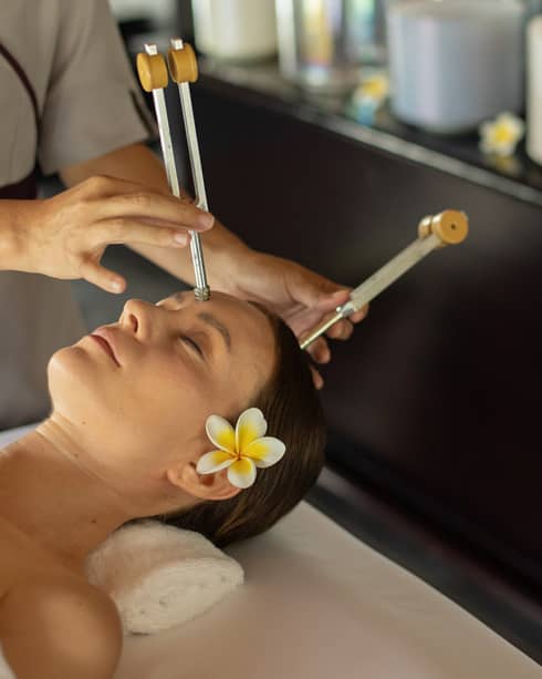 A practitioner delicately applies ohm tuning forks to the forehead and crown of a guest resting peacefully on a massage bed.