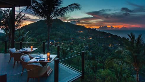 Corner of extended balcony with dining tables and chairs at dusk, expanse of forest, palm trees