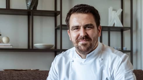 Smiling, bearded man wearing white chef's coat poses for camera while sitting on couch