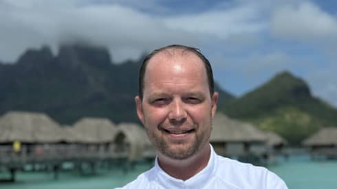 Smiling chef in white coat stands with arms crossed against backdrop of overwater pavilions