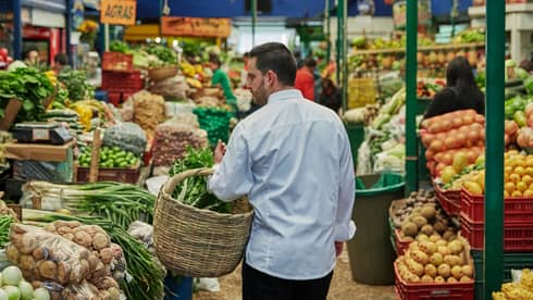 Back of man with beard carrying wicker basket through fruit market aisle, produce stacked on shelves