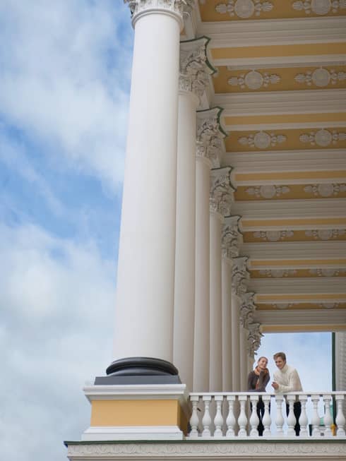 Looking up at man and woman peering over white corner balcony under tall white pillars, gold ornate ceiling