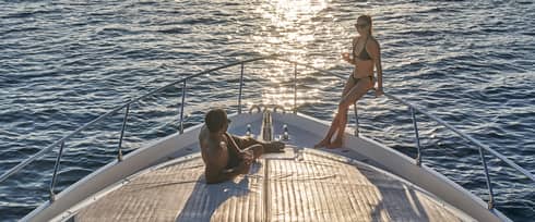 A couple in swimwear relaxing on the bow of a boat, wine glasses in hand and the setting sun glinting off the endless ocean.