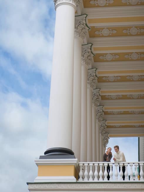 Looking up at man and woman peering over white corner balcony under tall white pillars, gold ornate ceiling