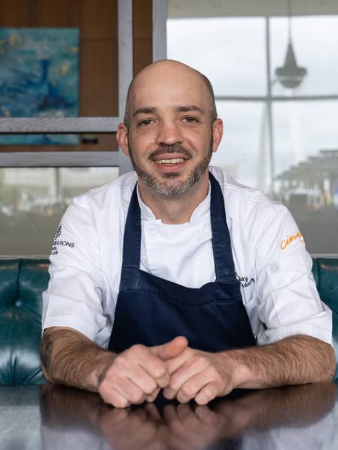 A bald male chef in a blue apron sitting at a table and booth.