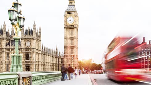 Red double-decker bus speeds by, Big Ben in the background as tourists stand nearby 