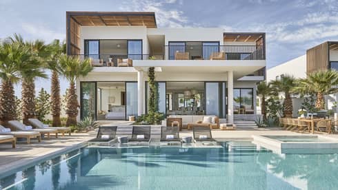 Modern luxury villa with a spacious pool area, surrounded by palm trees, featuring outdoor lounge chairs, a dining area, and floor-to-ceiling windows.