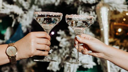 Two hands clink frosted-rimmed martini glasses in front of frosted Christmas tree