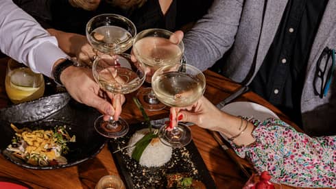 Four people toast with coupe glasses over a wooden table filled with small plates of food