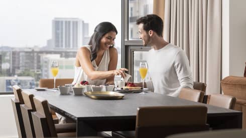 Smiling couple enjoys breakfast in their guest room, views of downtown Houston behind them 