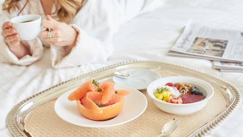 In-room dining tray with grapefruit, granola bowl, woman in bathrobe with coffee