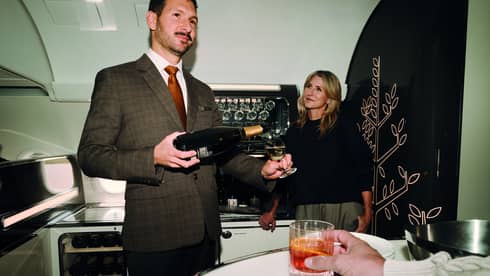 Person wearing brown tweed suit, white shirt and red tie pours a glass of wine for a private jet traveller standing nearby