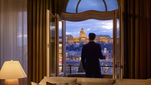 A person standing on a balcony at dusk, holding a glass of champagne, overlooking a lit historic building.