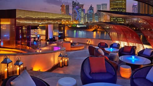 Rooftop bar furnished with round tables, plush chairs, lanterns, a curved bar area and a city skyline in the background