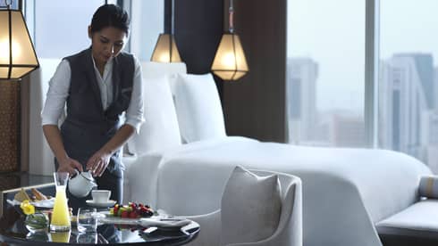 Woman in hotel suite pours tea into a white teacup next to a plate of fruit and bread.