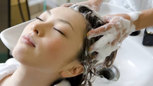 Woman closes eyes and leans back against sink as hands massage shampoo into her scalp