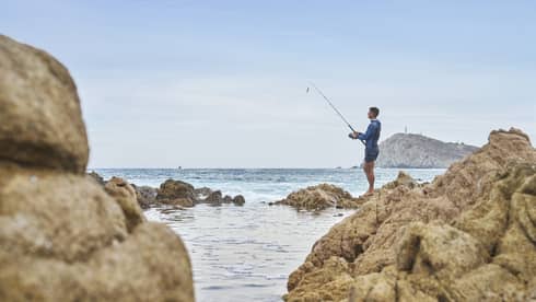 A fisher holding a pole stands barefoot atop a rocky outcrop and looks out toward an expanse of blue water under a clear sky.