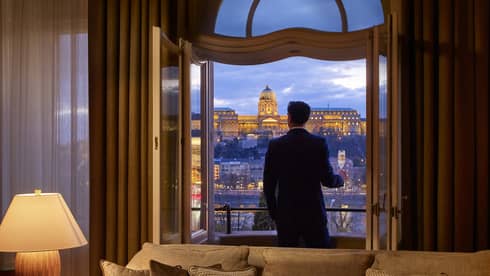 A person standing on a balcony at dusk, holding a glass of champagne, overlooking a lit historic building.
