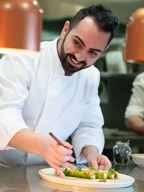 A chef with a small beard and moustache prepares a cannelloni with a green wrap and white sauce, on a white platter.