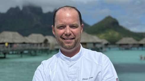 Smiling chef in white coat stands with arms crossed against backdrop of overwater pavilions