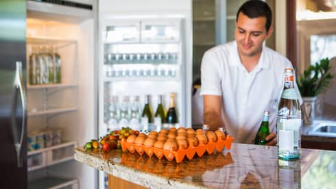 Smiling man holds green bottle in front of open fridge, fresh eggs on counter