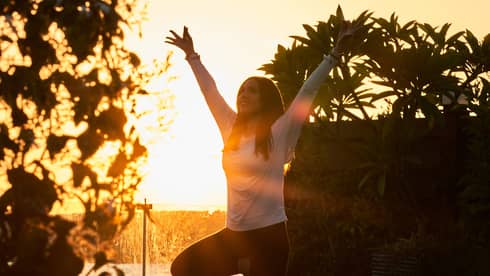 A person practising yoga in a tree pose at sunrise, surrounded by greenery with the sun shining brightly in the background.