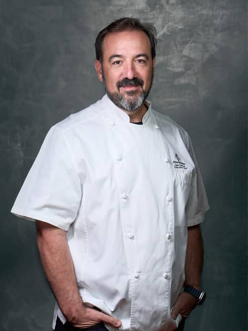 Smiling person in white double-breasted chef?s coat stands in front of gray background with hands in pockets