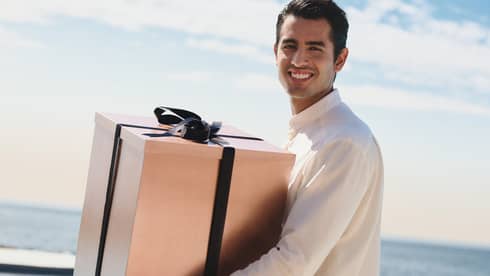 Man wearing white shirt and striped pants carries large present, with the ocean in the background