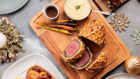 Overhead view of an assortment of dishes including a sliced beef Wellington, whole-roasted fish, yulie log and other desserts all on a light-grey table