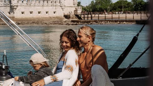 Two adults and a child relaxing on a sailboat near a historic stone tower on a sunny day.