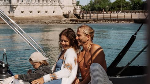 Two adults and a child relaxing on a sailboat near a historic stone tower on a sunny day.