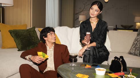 Two people sitting on a sofa with drinks, surrounded by snacks and game cards, enjoying a casual gathering in a stylish living room