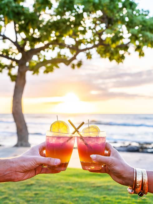 Two hands holding cocktails raised in a toast, the Maui sunset and North Pacific Ocean in the background