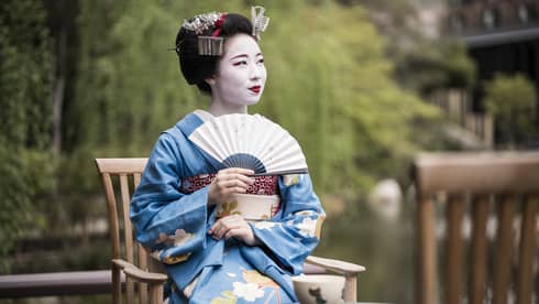 Geiko, a traditional Japanese entertainer, dressed in kimono and holding fan, sitting with cup of tea in garden