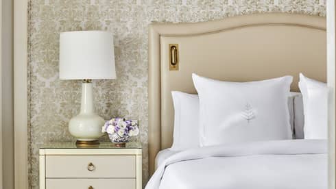 Guest bedroom with king bed, white linens and cream tufted headboard, foot bench, three-drawer side table with lamp, paisley wallpaper