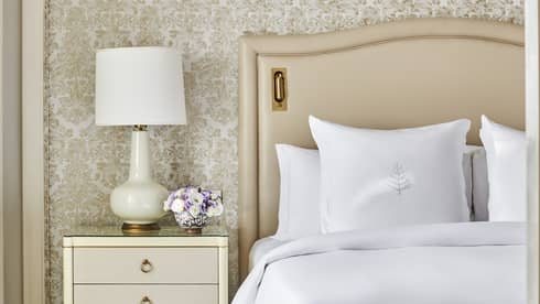 Guest bedroom with king bed, white linens and cream tufted headboard, foot bench, three-drawer side table with lamp, paisley wallpaper