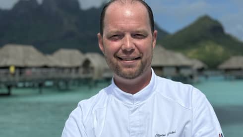 Smiling chef in white coat stands with arms crossed against backdrop of overwater pavilions