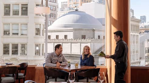 A four seasons staff takes a couple's order as they sit at a window table overlooking the city 