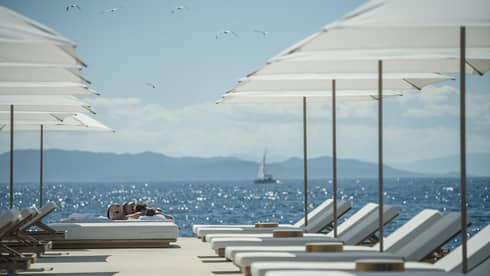 A sail boat sails in the distance while white lounge chairs are ready for guests and lined up next to the oceanside Nafsika Pool in Athens. 