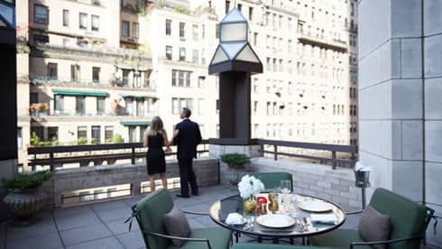 Man and woman in business suits look out from rooftop patio with table, plush chairs