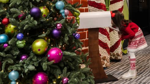 A child peers into white mailbox near a red and white striped archway that?s part of a large holiday display. A Christmas tree with colorful ornaments is in the foreground.