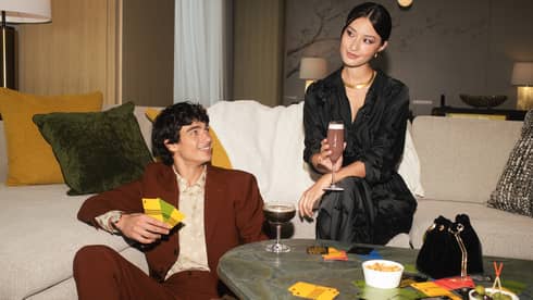 Two people sitting on a sofa with drinks, surrounded by snacks and game cards, enjoying a casual gathering in a stylish living room