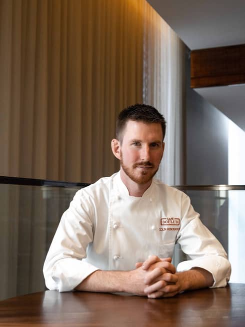 Chef de Cuisine Colin Henderson sitting at a wooden table in his chef whites