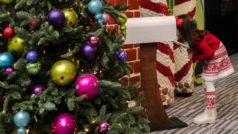 A child peers into white mailbox near a red and white striped archway that?s part of a large holiday display. A Christmas tree with colorful ornaments is in the foreground.